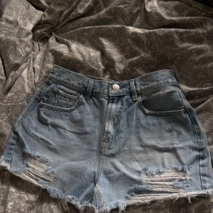 Ljusblå jeansshorts med slitningar - Snygga ljusblå jeansshorts med råa kanter och slitna detaljer framtill. Klassisk femficksmodell med knapp och dragkedja. Storlek M