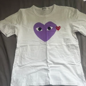 Vit t-shirt med lila hjärta från Comme des Garçons PLAY - Vit t-shirt från Comme des Garçons PLAY med stort lila hjärta och ögon tryckt på bröstet samt ett litet rött hjärta broderat bredvid. Klassisk rund halsringning och korta ärmar. Perfekt statement-plagg för dig som gillar streetwear.