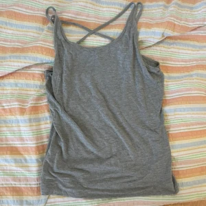 Grå linnetopp med korsade axelband - Säljer en enkel och stilren grå linnetopp med smala axelband som korsas i ryggen. Perfekt för varma dagar eller som lager under andra plagg. Mjuk och skön bomullsblandning.