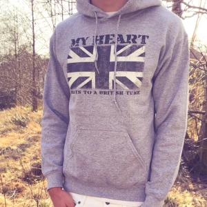 Grå hoodie från Drakenberg - Grå hoodie med tryck av brittiska flaggan och texten 'MY HEART BEATS TO A BRITISH TUNE' på bröstet. Tröjan har huva och magficka, samt ribbade muddar vid ärmslut och nederkant. Perfekt för en avslappnad stil.