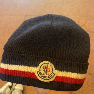 Mörkblå mössa från Moncler med röd och vit rand - Snygg mörkblå Moncler-mössa med ribbstickat material och klassiska ränder i rött och vitt runt om. Framtill sitter Monclers logotyp. Perfekt accessoar för kyliga dagar. Äkta 