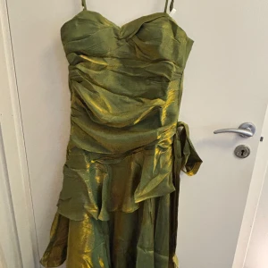 Grön guldig axelbandsklänning med omlott och volang - Säljer en glansig grön klänning med guldskimmer och smala axelband. Klänningen har omlott-detaljer, volanger och knyt i midjan. Perfekt för festliga tillfällen och har en elegant, figurnära passform.