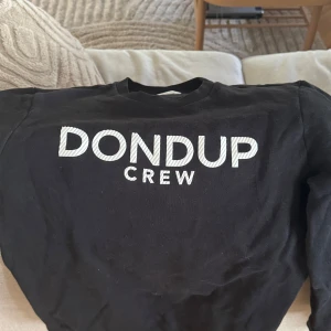 Svart sweatshirt från Dondup - Säljer en svart sweatshirt från Dondup med vit text 'DONDUP CREW' på bröstet. Tröjan har rund hals och långa ärmar, perfekt för en avslappnad stil. Passar dig som gillar klassiska och stilrena plagg. Storlek liknar M
