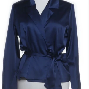 Mörkblå omlottjacka med knytband - Snygg mörkblå omlottjacka med krage och knytband i midjan. Jackan har lång ärm och en elegant, lite glansig finish som ger en stilren look. Perfekt för dig som vill ha en klassisk men trendig jacka. Det är strl M men den är mer som en S