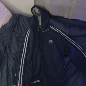 Mörkblå hoodiejacka från Lonsdale - Mörkblå hoodiejacka i tjockt fleece tyg från Lonsdale med dragkedja och vita ränder längs ärmarna. Jackan har huva med ljusgrå insida och broderad logga på bröstet. Perfekt för en sportig stil.