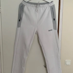 Boss Hadim byxor - M - Snygga vita mjukisbyxor från BOSS med grå paneler vid fickorna och midjan. Byxorna har dragsko i midjan, två dragkedjefickor fram och BOSS-logga på benet. Perfekta för en avslappnad stil.
