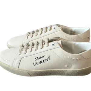 Vita sneakers från Saint Laurent - Säljer ett par stilrena vita sneakers från Saint Laurent med handskriven logga på sidan. Skorna har snörning och en platt sula, perfekt för en clean och modern look. De är ser nya ut och jag säljer pga att de är för små