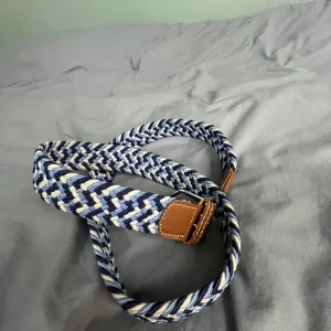 Blått flätat bälte med skinn-detaljer - Säljer ett snyggt flätat bälte i blått, vitt och grått med bruna skinn-detaljer vid spännet. Perfekt accessoar för att lyfta din outfit med en sportig och stilren touch. Bältet har ett flexibelt material och passar till många olika looks.