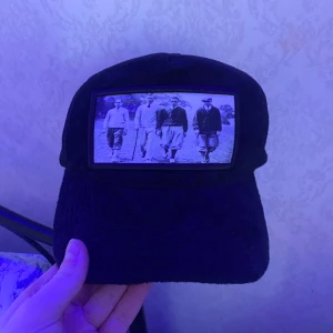 Svart keps med vintagefoto - Svart keps med mesh på sidorna och ett stort tygmärke framtill med ett svartvitt vintagefoto av fyra personer som går tillsammans. Kepsen har böjd skärm och justerbar passform bak.