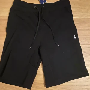 Svarta shorts från Polo Ralph Lauren - Svarta shorts från Polo Ralph Lauren med smal passform och snörning i midjan. Klassisk broderad logga på ena benet och bakficka. Perfekta för sommaren och tillverkade i mjuk bomull.