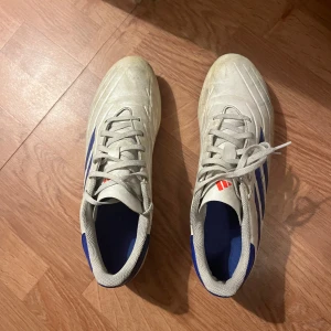 Adidas Copa fotbollsskor - Säljer ett par Adidas Copa fotbollsskor i vitt med blå och röda detaljer. Skorna har klassiska tre ränder på sidan och snörning framtill. Yttermaterialet är syntet och sulan har fasta dobbar för spel på gräs. Ställ frågor😁.