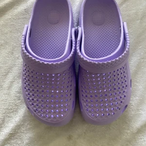 Ljuslila tofflor  - Säljer ett par ljuslila tofflor med klassisk design och perforerad ovandel för bra ventilation. Skorna har en justerbar rem bak och är tillverkade i syntetmaterial. Perfekta för sommaren eller som inneskor. Stlk kvar 35-36 och 36-37 , 89kr/ par , lila färg 