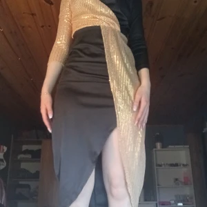 Svart och guldig långklänning med paljettdetaljer - Säljer en unik långklänning i svart med guldiga paljetter som sveper över axeln och längs sidan. Klänningen har lång ärm, samt en hög slits framtill. Perfekt för dig som vill sticka ut!