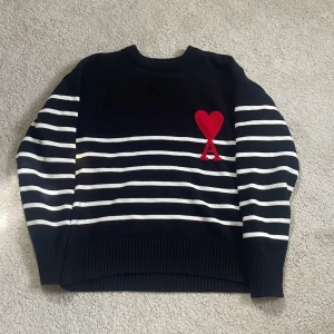 Svart och vit randig stickad tröja från Ami Paris - Säljer en svart stickad tröja med vita horisontella ränder och ett rött hjärta med bokstaven A broderat på bröstet. Tröjan har rund hals och långa ärmar, perfekt för en stilren och trendig look.