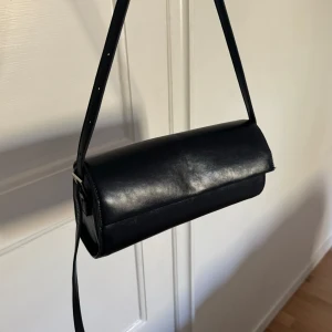 Svart cylindrisk axelväska - Säljer en stilren svart axelväska med enkel design och justerbart axelband. Väskan har ett avlångt format. Perfekt för dig som gillar minimalistisk stil.