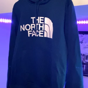 Blå hoodie från The North Face - Säljer en blå hoodie från The North Face med stor vit logga på bröstet. Tröjan har huva med dragsko och en klassisk känguruficka framtill. Perfekt för en avslappnad och sportig stil.