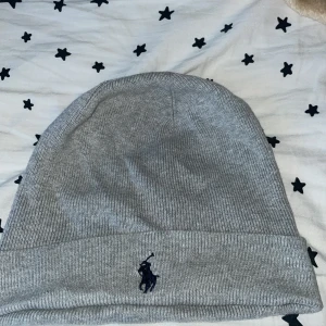 Grå ribbad mössa från Polo Ralph Lauren - Snygg grå ribbstickad mössa från Polo Ralph Lauren med klassisk broderad logga framtill. Perfekt för kyliga dagar och enkel att matcha med olika outfits. Mjuk och bekväm passform.