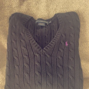 Mörkgrå kabelstickad tröja från Ralph Lauren Sport - Kabelstickad mörkgrå tröja från Ralph Lauren Sport med v-ringad hals och lila broderad logga på bröstet. Tillverkad i 100% bomull och har långa ärmar. Klassisk och stilren design som passar till mycket. Använd en del men är i fint skick! 