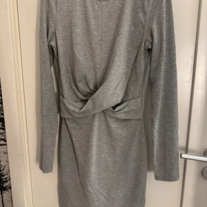 Grå omlottklänning med lång ärm - Säljer en stilren grå klänning med omlottdetalj framtill och långa ärmar. Klänningen har en kroppsnära passform och är tillverkad i ett mjukt, stretchigt material som känns bekvämt mot huden. Perfekt för dig som gillar enkel men snygg stil.