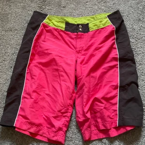 Färgglada shorts från Index - Shorts i stark rosa färg med svarta sidor och limegrön linning. De har vita detaljer längs sidorna och knäppning framtill. Ganska baggy och tunt material , skriv för mått