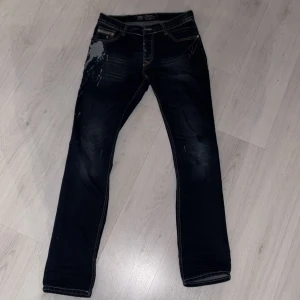 Mörkblå jeansbyxor från Diesel - Säljer ett par mörkblå jeansbyxor från Diesel med slitningar och unika detaljer på fickorna. Byxorna har raka ben och klassisk femficksdesign. Snygga kontrastsömmar och en cool tvättad look.
