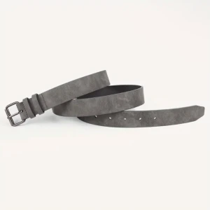 Grått mockabälte med silverfärgat spänne - Snyggt grått bälte i mocka med klassiskt silverfärgat spänne. Bältet har en stilren design och flera hål för justerbar passform. Perfekt accessoar för att lyfta din outfit.