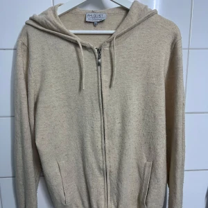  Kashmir tröja från Ahlquist Clothing - Mysig beige hoodie från Ahlquist Clothing i storlek M. Tröjan har dragkedja framtill, huva med snören och två fickor. Perfekt för en avslappnad stil och enkel att matcha med det mesta.