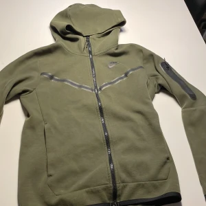 Grön  niketech - Säljer en grön hoodiejacka från Nike med hel dragkedja och huva. Jackan har svarta detaljer, en diskret Nike-logga på bröstet och en ficka med dragkedja på ena ärmen. Perfekt för en sportig stil.