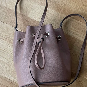 Rosa bucket bag med axelrem - Säljer en snygg rosa bucket bag med justerbar axelrem och dragskostängning. Väskan har en stilren design med silverfärgade metalldetaljer och är tillverkad i syntetmaterial. Perfekt för dig som vill ha en trendig och praktisk accessoar. En mini väska