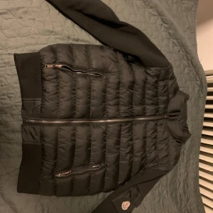Svart cardigan från Moncler - Säljer en svart cardigan från Moncler i storlek M. Jackan har långa ärmar, två snedställda fickor med dragkedja och Moncler-logga på ärmen. Klassisk quiltad design och stängs med dragkedja framtill.