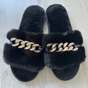 Svarta fluffiga sandaler med guldkedja - Säljer ett par svarta sandaler med mjuk, fluffig päls och en glittrig guldfärgad kedjedetalj över foten. Perfekta för att ge din outfit en lyxig touch. Snygga och trendiga att bära både hemma och ute. Det passar storlek 40-41 Det är helt nya och aldrig använda. Kan säljas för 150 kr vid snabb affär.