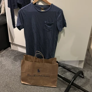 Blå t-shirt från Polo Ralph Lauren - Säljer en stilren blå t-shirt från Polo Ralph Lauren med rund halsringning och en liten bröstficka. Perfekt basplagg med klassisk passform och diskret logga på insidan av kragen.