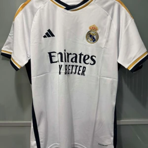 Real Madrid hemmatröja  - Säljer en vit Real Madrid fotbollströja från Adidas med klubbmärke och sponsortryck på bröstet. Tröjan har guld- och marinblå detaljer längs axlar och sidor samt korta ärmar. Materialet är lätt och andas, perfekt för match eller träning. Finns alla storlekar 