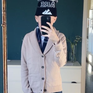 Beige cardigan - Säljer en beige kofta med sjalkrage och stora knappar framtill. Tröjan har lång ärm och ribbade muddar vid ärmslut och nederkant. Perfekt för lager-på-lager och enkel att matcha med olika stilar.