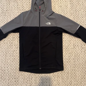 North face windbreaker - Säljer en snygg svart och grå vindjacka från The North Face med huva och hel dragkedja. Jackan har två sidofickor och logga på bröstet. Perfekt för blåsiga dagar och enkel att matcha med olika outfits.