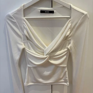 Vit långärmad topp med twist framtill från Bik Bok - Trendig vit långärmad topp från Bik Bok med snygg twistdetalj framtill och djup v-ringning. Toppen är figurnära och har en croppad passform som passar perfekt till både jeans och kjol.