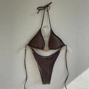 Brun bikini med gulddetaljer - Säljer en brun bikini från Shein i storlek M. Bikinin har triangeltopp med knyt i nacken och dekorativa guldfärgade kedjedetaljer på båda sidor av överdelen. Passar S/M☀️🤎