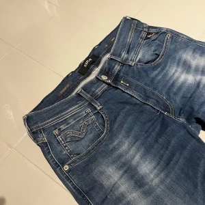Replay Anbass jeans i blå denim - Säljer ett par Replay Anbass jeans i klassisk blå tvätt med snygga slitningar. Jeansen har fem fickor, raka ben och detaljerade sömmar på bakfickan. Passformen är normal och materialet är mjuk denim med stretch. Midjemått W33 och längd L32. HELT NYA!!!