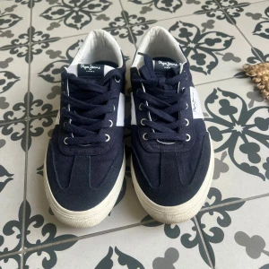 Mörkblå sneakers från Pepe Jeans - Snygga mörkblå sneakers från Pepe Jeans med vita detaljer och vit sula. Skorna har snörning och en klassisk låg modell med rund tå. Perfekta för en avslappnad stil och passar till det mesta.