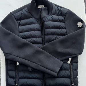 Moncler Cardigan Svart - Svart Moncler-cardigan i stilren design med en mix av stickat tyg och tekniska detaljer. Försedd med dragkedja och ribbade muddar för en smal passform. Det klassiska Moncler-logotypen pryder ärmen, vilket ger en lyxig men diskret touch.