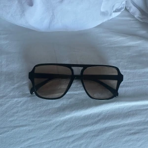 Svarta solglasögon med fyrkantig form - Stilrena svarta solglasögon med markerad fyrkantig båge och mörka glas. Bågarna är breda och har en modern, oversized känsla. Perfekta för dig som vill ha en cool och tidlös accessoar.