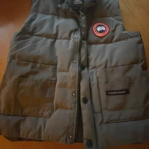 Grå dunväst från Canada Goose - Snygg grå dunväst från Canada Goose med klassisk logga på bröstet och stora fickor framtill. Västen har knappar och är perfekt för lager-på-lager-stil under lite kyligare dagar.                                                                                                                                   Vill du veta något mer eller vill ha fler bilder så är det bara att skriva! Priset är såklart inte hugget i sten!