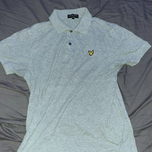 Ljusgrå pikétröja från Lyle & Scott - Säljer en ljusgrå pikétröja från Lyle & Scott med klassisk krage och två knappar framtill. Tröjan har korta ärmar och den ikoniska gula loggan broderad på bröstet. Perfekt för en stilren och avslappnad look.
