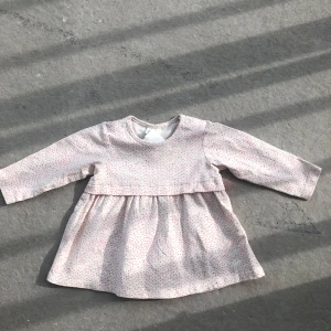 Vit klänning med rosa och gröna prickar - Supersöt vit klänning med lång ärm och små rosa samt gröna prickar över hela tyget. Klänningen har en lätt utställd kjol och tryckknapp vid axeln för enkel påklädning. Perfekt för vårens och sommarens dagar.