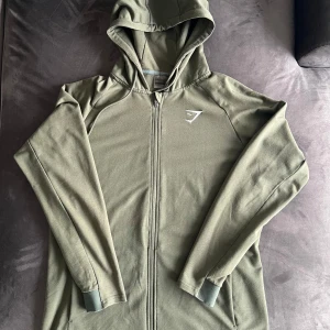 Olivgrön zip up Gymshark - Säljer en olivgrön hoodie från Gymshark med hel dragkedja och huva. Har en smickrande passform som är slimfit. Storlek XL