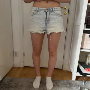 Ljusblå jeansshorts med fransar - Säljer ett par ljusblå jeansshorts med råa franskantade benslut och klassisk femficksdesign. Shortsen har normal passform och knappgylf. Perfekta för varma dagar!