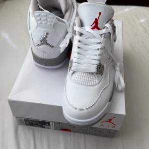 Nike Air Jordan 4 White Oreo - Säljer ett par fräscha, sprillans nya Nike Air Jordan 4 White Oreo med vit ovandel i skinn, grå detaljer och röd Jumpman-logga på plösen. Klassisk siluett med snörning och meshpaneler på sidorna. Perfekta för dig som gillar stilrena sneakers. 