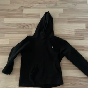 Svart hoodie från Polo Ralph Lauren - Säljer en svart hoodie från Polo Ralph Lauren med klassisk huva och magficka. Tröjan har en liten broderad logga på bröstet och är tillverkad i mjukt material. Perfekt för en avslappnad och stilren look.