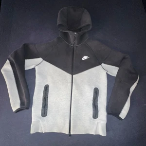 Nike tech fleece - Säljer en stilren svart och grå hoodie från Nike med hel dragkedja och huva. Jackan har två fickor framtill med svarta detaljer och en vit Nike-logga på bröstet. Perfekt för en sportig look.