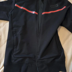 Svart sportjacka med röd rand från Brando - Svart sportig jacka från Brando med dragkedja framtill och röda ränder över bröstet. Jackan har ribbade muddar och en stilren design som passar perfekt till träning eller vardagslooken.